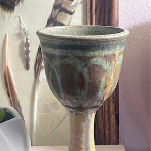 May include: A ceramic goblet featuring a flared rim and a tall, slender stem. The bowl displays a mottled glaze in shades of deep brown, forest green, and muted teal, with drips of glaze running down the stem onto the textured, light tan base.