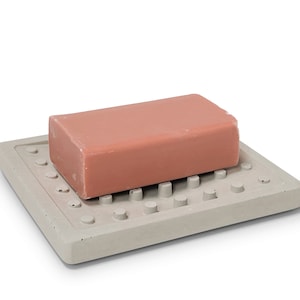 Può includere: Un portasapone in cemento grigio chiaro con una griglia di piccoli cerchi in rilievo. Una saponetta rosa poggia sul portasapone.