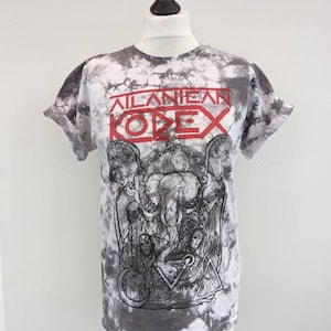 Puede incluir: Una camiseta gris y blanca con efecto tie-dye y mangas enrolladas. La camiseta presenta el texto rojo "ATLANTEAN KODEX" sobre un gráfico detallado en blanco y negro de un elefante y otras figuras. La camiseta está en un maniquí.