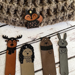 May include: Five leather animal tags featuring different woodland creatures. The tags are brown, grey, and tan with black details. The animals include a fox, a deer, a bear, a hedgehog, and a rabbit.