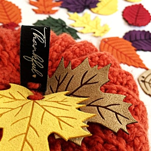 May include: A red knitted pumpkin with a gold tag that says "Thankful" and is decorated with felt leaves in shades of yellow, brown, and beige.