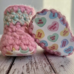 May include: Pink crocheted baby booties with a white fabric lining featuring a pattern of candy hearts with various sayings, such as "Be Mine", "Love Bug", "Dream Big", "Kiss Me", "Hug Me", "Sweet Heart", and "Puppy Love".