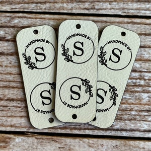 Peut inclure: Trois étiquettes en cuir blanc cassé avec un trou en haut. Chaque étiquette a un cercle noir avec une lettre noire "S" à l'intérieur et une couronne noire autour du cercle. Le texte "HANDMADE BY" est écrit autour du cercle.
