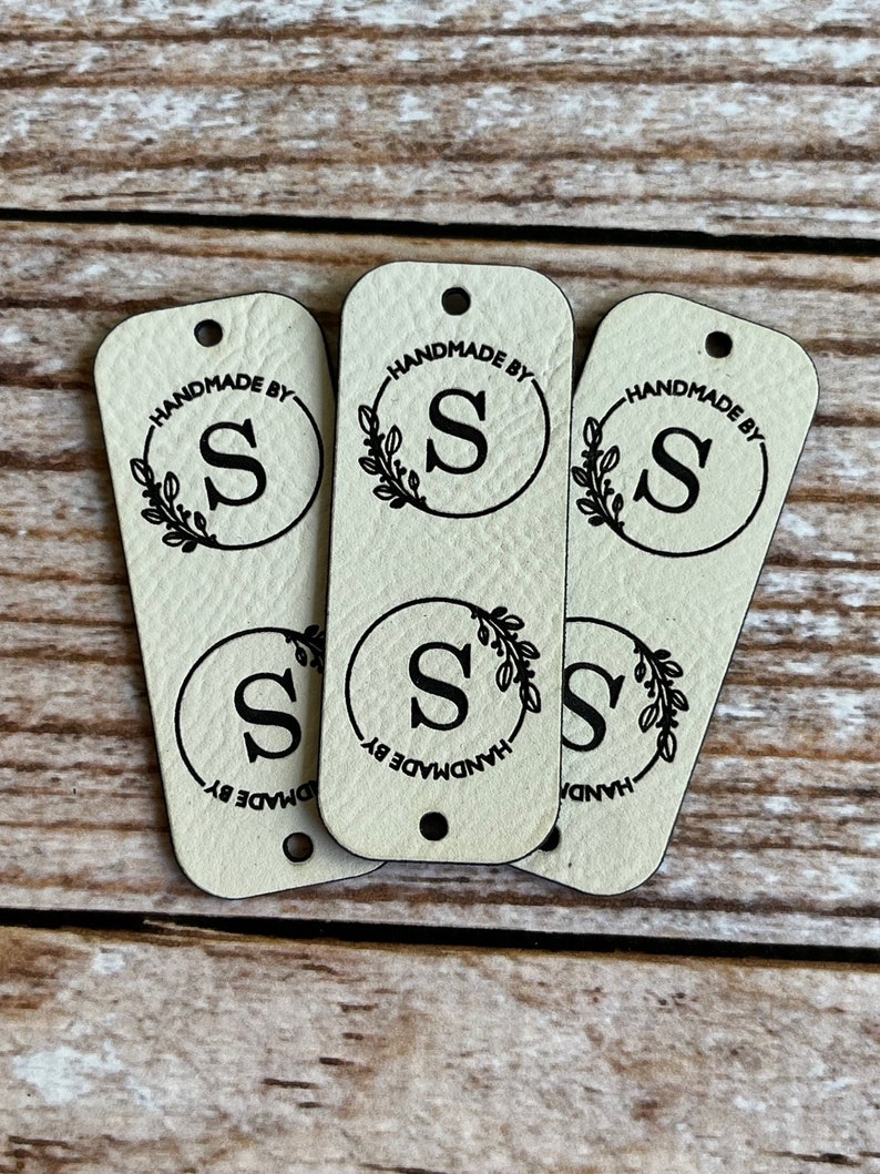 Pode incluir: Tr&ecirc;s etiquetas de couro bege com um furo no topo. Cada etiqueta tem um c&iacute;rculo preto com uma letra preta "S" dentro e uma coroa preta ao redor do c&iacute;rculo. O texto "HANDMADE BY" est&aacute; escrito ao redor do c&iacute;rculo.