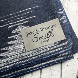 Peut inclure: Une couverture en laine bleu marine et blanche à motifs avec une étiquette en cuir sur laquelle est inscrit "Jake & Breanne Smith 6.18.24".