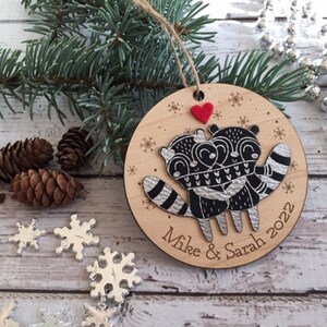 May include: A round wooden Christmas bauble featuring a black and white illustration of two raccoons embracing. The bauble is decorated with snowflakes and a red heart. The text "Mike & Sarah 2022" is inscribed on the bottom of the bauble.
