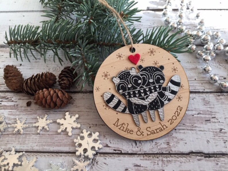 May include: A round wooden Christmas bauble featuring a black and white illustration of two raccoons embracing. The bauble is decorated with snowflakes and a red heart. The text "Mike & Sarah 2022" is inscribed on the bottom of the bauble.
