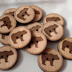 May include: A collection of wooden buttons featuring a bear silhouette design. The buttons are light brown with a dark brown bear silhouette. The buttons are approximately 1 inch in diameter.