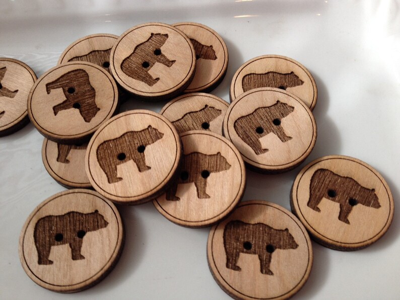 May include: A collection of wooden buttons featuring a bear silhouette design. The buttons are light brown with a dark brown bear silhouette. The buttons are approximately 1 inch in diameter.