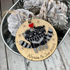 May include: A wooden Christmas bauble featuring two raccoons embracing, a red heart, and the inscription "Alyssa & Joe 2024".