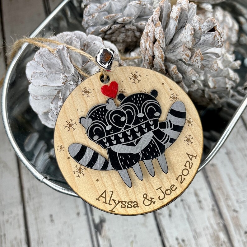 May include: A wooden Christmas bauble featuring two raccoons embracing, a red heart, and the inscription "Alyssa & Joe 2024".