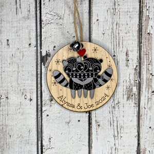 May include: A round wooden bauble featuring a pair of raccoons embracing, a red heart, and snowflakes. The bauble is personalised with the text "Alyssa & Joe 2024".