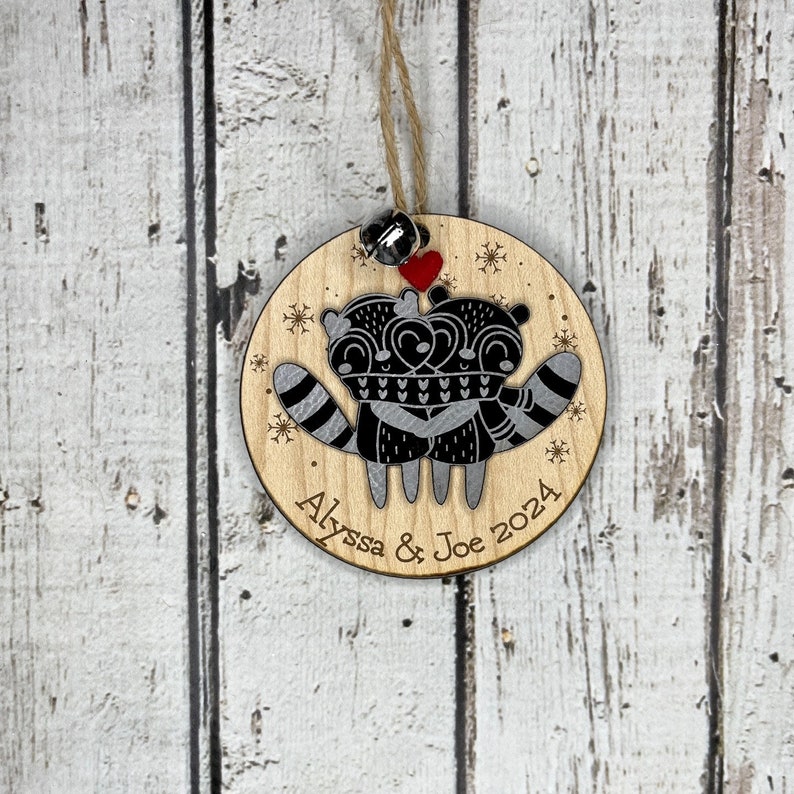 May include: A round wooden bauble featuring a pair of raccoons embracing, a red heart, and snowflakes. The bauble is personalised with the text "Alyssa & Joe 2024".