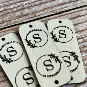 Pode incluir: Quatro etiquetas de couro bege com letras pretas. Cada etiqueta tem um c&iacute;rculo com a letra "S" dentro e as palavras "Handmade by" ao redor do c&iacute;rculo. O c&iacute;rculo &eacute; cercado por uma coroa de folhas.