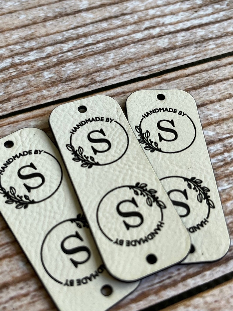 Pode incluir: Quatro etiquetas de couro bege com letras pretas. Cada etiqueta tem um c&iacute;rculo com a letra "S" dentro e as palavras "Handmade by" ao redor do c&iacute;rculo. O c&iacute;rculo &eacute; cercado por uma coroa de folhas.