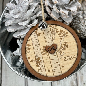 May include: A round wooden ornament with a birch tree design, a heart with the initials "C+R" and the year "2024" engraved on it. The ornament has a brown border and a silver bell.