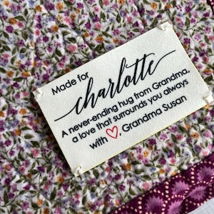May include: A white tag with black text that reads "Made for Charlotte A never-ending hug from Grandma, a love that surrounds you always with Grandma Susan". The tag is attached to a white fabric with a floral pattern.