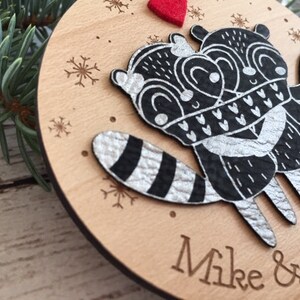 May include: A wooden Christmas bauble featuring two black and white raccoons embracing, with silver glitter details. The bauble is personalised with the names "Mike & Sarah".