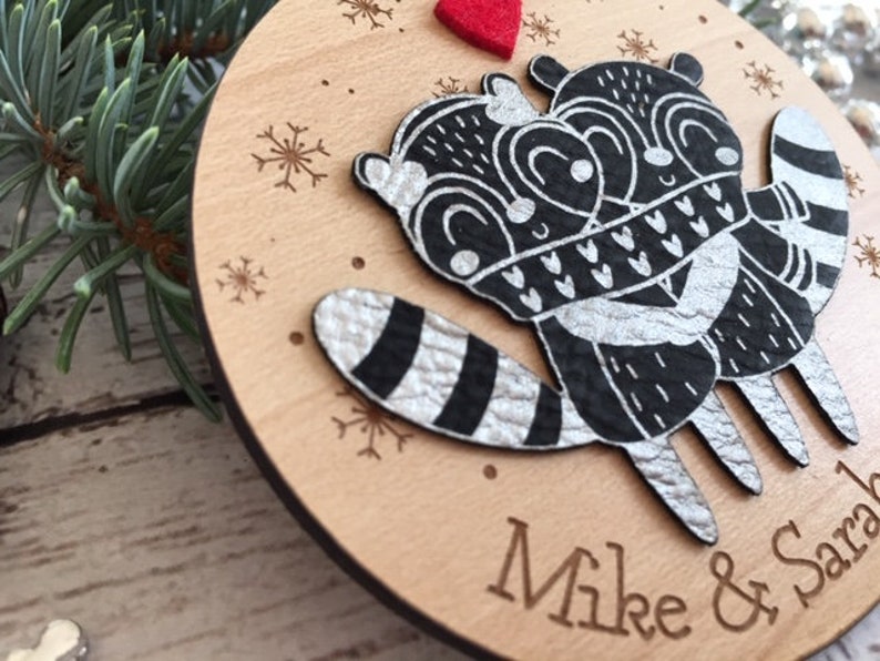 May include: A wooden Christmas bauble featuring two black and white raccoons embracing, with silver glitter details. The bauble is personalised with the names "Mike & Sarah".
