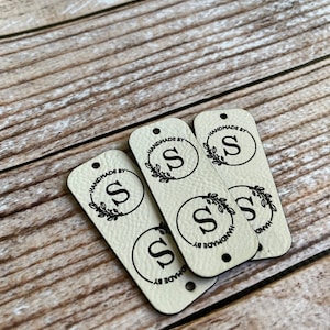 Pode incluir: Quatro etiquetas de couro bege com um design circular preto. Cada c&iacute;rculo tem uma letra preta "S" e um design de folhas pretas. O texto "Handmade by" &eacute; impresso em torno do c&iacute;rculo em cada etiqueta.