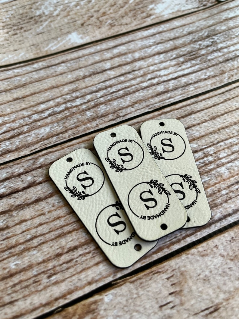 Pode incluir: Quatro etiquetas de couro bege com um design circular preto. Cada c&iacute;rculo tem uma letra preta "S" e um design de folhas pretas. O texto "Handmade by" &eacute; impresso em torno do c&iacute;rculo em cada etiqueta.
