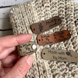 May include: Four brown leather labels with white text. The labels say "Made by Sarah", "Made by Sandra", "Made by Sandra", and "Made with by Mom". The labels have a small design of a leaf and a heart.