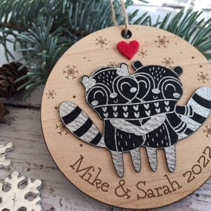 May include: A round wooden Christmas bauble featuring a black and white illustration of two raccoons embracing. The bauble is decorated with snowflakes and the text "Mike & Sarah 2022". A small red felt heart is attached to the top of the bauble.