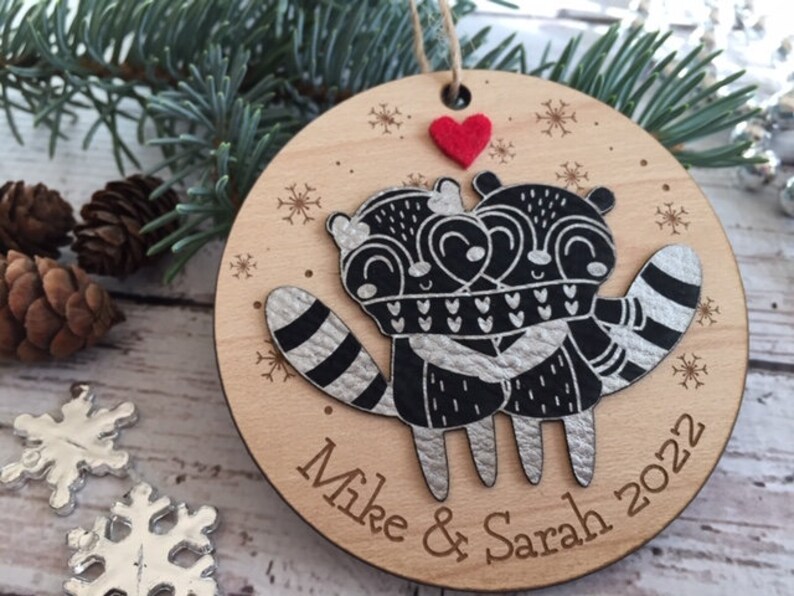 May include: A round wooden Christmas bauble featuring a black and white illustration of two raccoons embracing. The bauble is decorated with snowflakes and the text "Mike & Sarah 2022". A small red felt heart is attached to the top of the bauble.