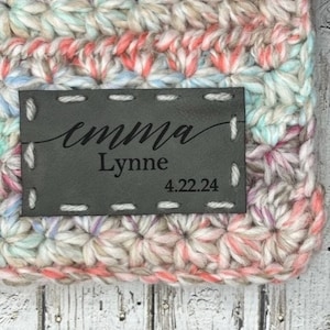 Peut inclure: Une étiquette en cuir gris avec le nom "emma Lynne" et la date "4.22.24" cousue avec du fil noir. L'étiquette est attachée à une couverture au crochet avec un motif multicolore.