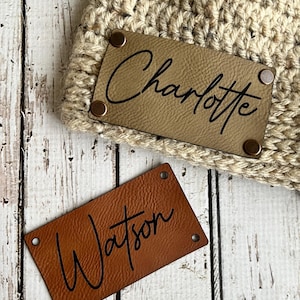 May include: Two personalized leather labels with the names "Charlotte" and "Watson" written in cursive. The labels are brown and have metal rivets around the edges.