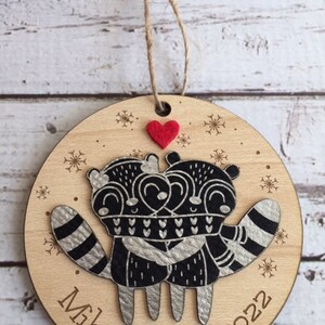 May include: A round wooden Christmas bauble featuring a pair of raccoons embracing. The raccoons are black and silver with white details. The bauble is personalised with the names "Mike & Sarah" and the year "2022".