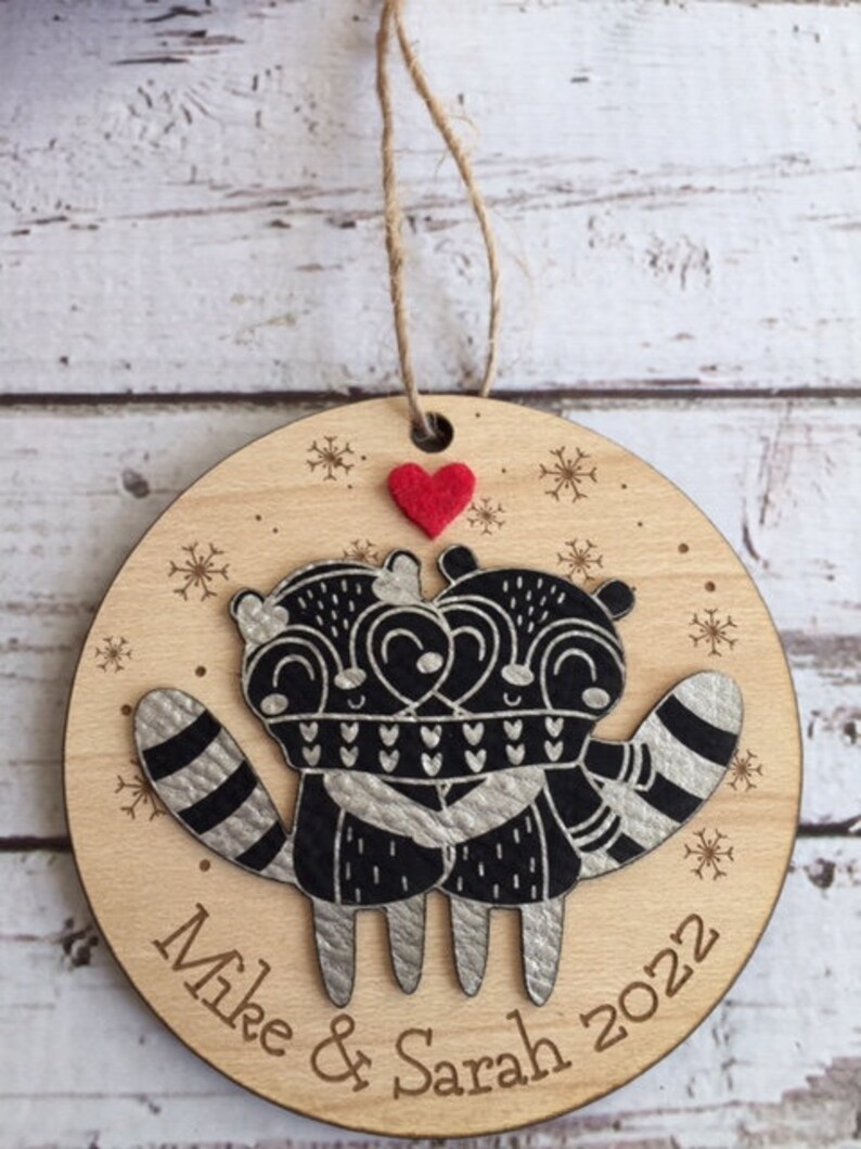 May include: A round wooden Christmas bauble featuring a pair of raccoons embracing. The raccoons are black and silver with white details. The bauble is personalised with the names "Mike & Sarah" and the year "2022".