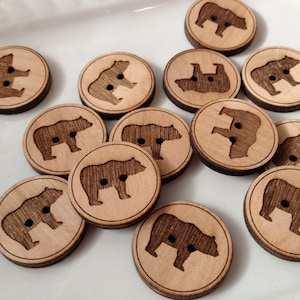 May include: A collection of wooden buttons featuring a bear silhouette design. The buttons are brown with a natural wood grain and have two holes for sewing.
