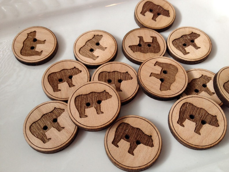 May include: A collection of wooden buttons featuring a bear silhouette design. The buttons are brown with a natural wood grain and have two holes for sewing.