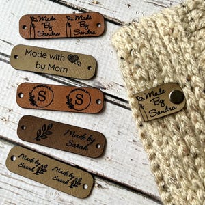 May include: Assortment of six brown leather labels with various text and designs. The labels include "Made by Sandra", "Made with by Mom", "Handmade by S", "Made by Sarah", and "Made by Sandra".