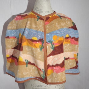 May include: A brown fleece cape with a colorful desert sunset print. The cape has a brown trim and a button closure at the neck.