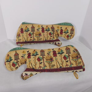 Puede incluir: Un par de guantes para horno con un estampado colorido de casas para pájaros. Los guantes están hechos de tela acolchada y tienen un lazo de colgar amarillo. El texto "Backyard Birdhouses" está impreso en la parte inferior de los guantes.