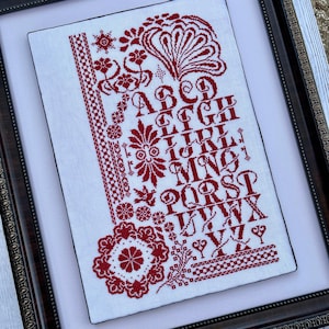 May include: A framed cross-stitch sampler with a red alphabet, floral designs, and a red square with a four-leaf clover in the bottom left corner.