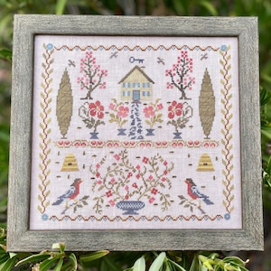 May include: A cross-stitch embroidery pattern featuring a small house with a key above it, two trees, and a beehive on either side. The house is surrounded by flowers and a vase of flowers. The pattern is framed in a gray frame.
