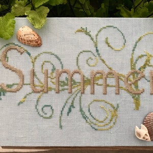 Op de afbeelding: Borduurwerk met kruissteek op een lichtblauw canvas met het woord "Summer" in bruin en groen garen. Het woord is omgeven door groene en gele bloemmotieven. Twee schelpen liggen op het canvas.