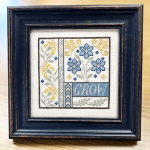 May include: Framed cross-stitch art with yellow and blue floral patterns. The artwork includes the word "GROW" in a blue banner. The frame is a distressed navy blue, giving a rustic feel to the piece.