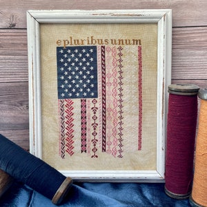 May include: A framed cross-stitch of an American flag with the words "e pluribus unum" above it. The flag is surrounded by a white frame and is sitting on a blue surface.