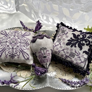 May include: Three lavender-themed embroidered pillows on a silver tray with a lace doily. The pillows are square and feature a purple and white floral design. The tray is decorated with lavender sprigs.