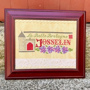 May include: A framed cross-stitch embroidery on cream fabric featuring a stone castle with a red door, the text La Belle Bretagne and JOSSELIN, and purple flowers. The piece is bordered by white lace trim and encased in a dark red wooden frame.