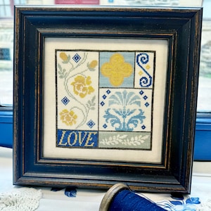 Peut inclure: Une œuvre d'art au point de croix encadrée dans un cadre bleu marine. L'œuvre présente un motif patchwork avec des motifs floraux et géométriques en jaune, bleu et blanc. Le mot "LOVE" est brodé en bleu en bas. Une bobine de fil bleu est au premier plan.