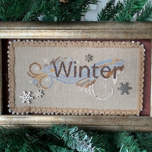 Op de afbeelding: Een ingelijst kruissteek met een beige achtergrond en het woord "Winter" in blauwe en bruine letters. Het ontwerp bevat gestileerde sneeuwvlokken en swirls. De lijst is goud met een bruine passe-partout.