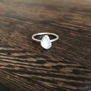May include: A silver ring featuring a pear-shaped, white gemstone. The band has a hammered texture. The ring is set against a dark wood grain background.
