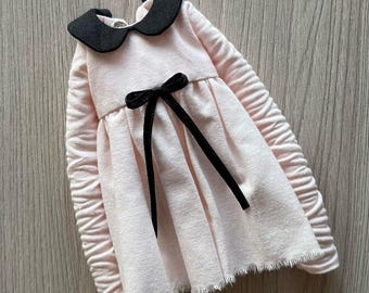 May include: A pink dress with a black peter pan collar and a black bow at the waist. The dress has long sleeves and a ruffled hem.