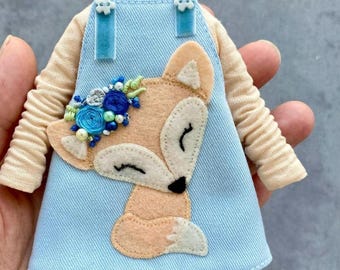 May include: A light blue denim overall dress with a fox applique. The fox is wearing a flower crown with blue and white flowers and pearls. The dress has blue velvet straps and white buttons.