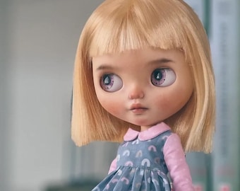 May include: A doll with blonde hair and bangs wearing a blue dress with a pink collar and rainbow pattern. The doll has large blue eyes with purple accents.